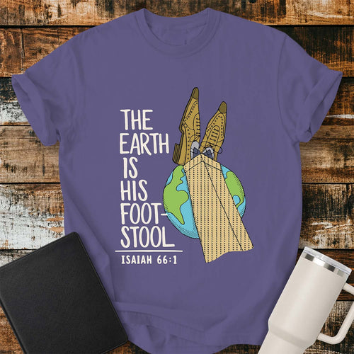 Grape purple T-shirt featuring a colorful illustration of the Earth with a pair of legs in brown dress shoes and tan herringbone trousers resting on top, visually representing the phrase “The Earth Is His Footstool.” Cream-colored stacked lettering reads, “THE EARTH IS HIS FOOTSTOOL,” with “ISAIAH 66:1” below. The shirt is displayed on a rustic wood plank background with a black Bible on the left and a white insulated tumbler on the right.