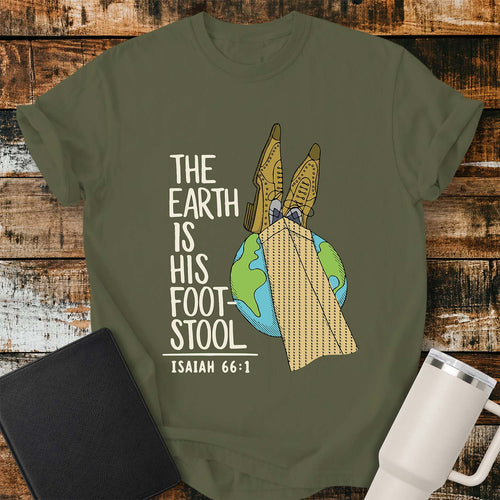 hemp green T-shirt featuring a colorful illustration of the Earth with a pair of legs in brown dress shoes and tan herringbone trousers resting on top, visually representing the phrase “The Earth Is His Footstool.” Cream-colored stacked lettering reads, “THE EARTH IS HIS FOOTSTOOL,” with “ISAIAH 66:1” below. The shirt is displayed on a rustic wood plank background with a black Bible on the left and a white insulated tumbler on the right.