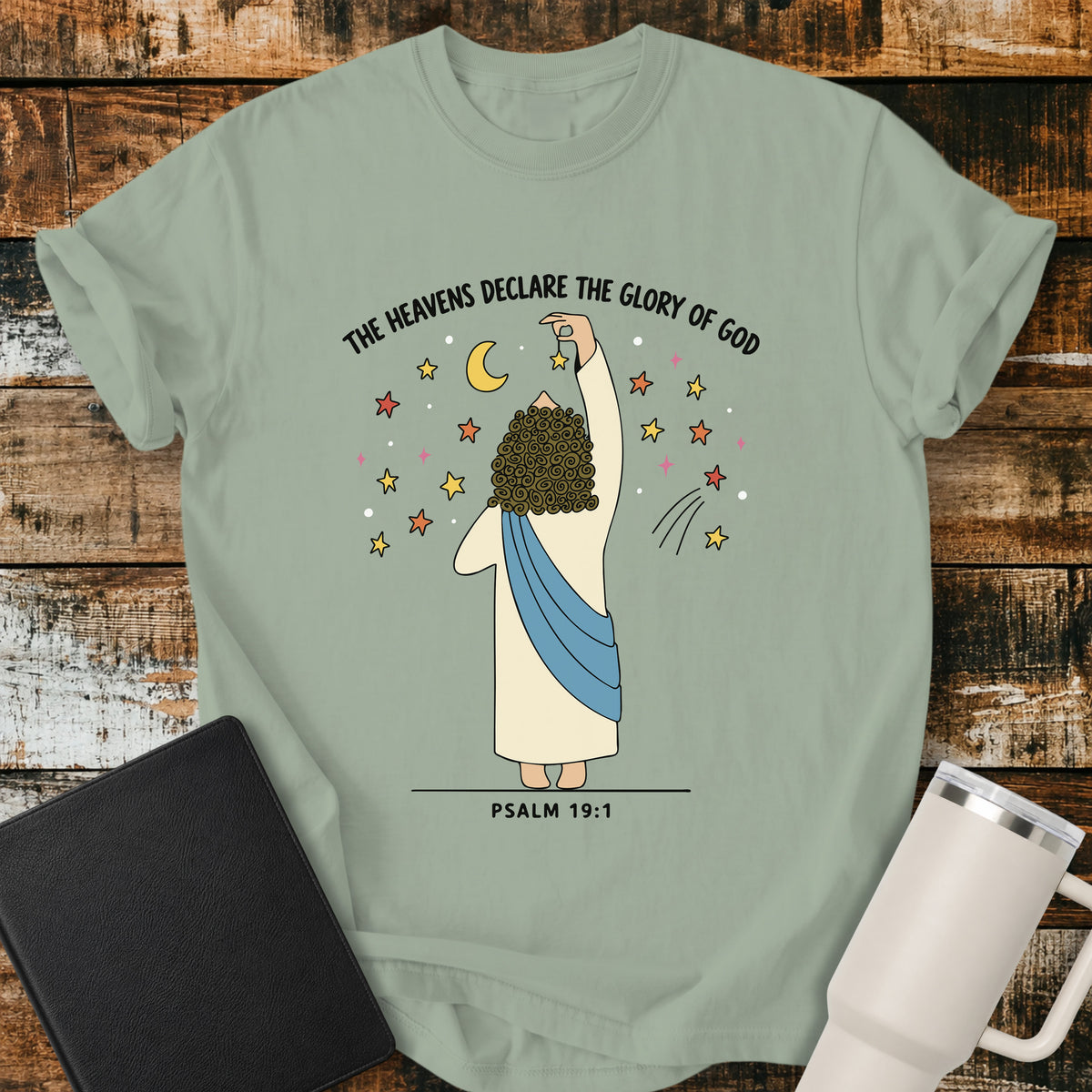 bay green Christian t-shirt featuring a whimsical illustration of God placing stars in the heavens, with the text “The Heavens Declare the Glory of God” and Psalm 19:1.