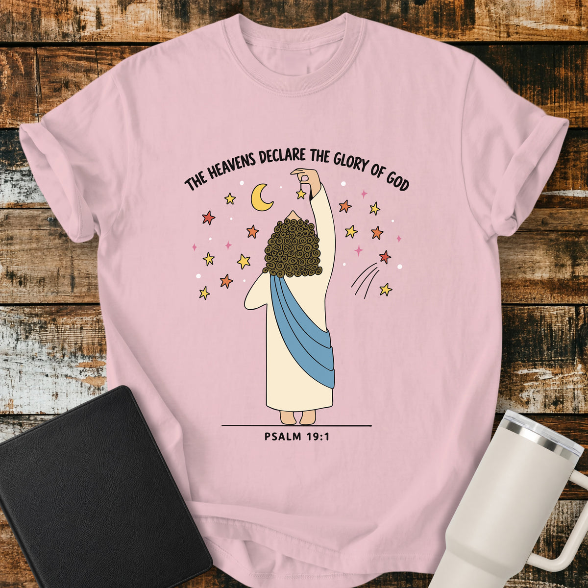 blossom pink Christian t-shirt featuring a whimsical illustration of God placing stars in the heavens, with the text “The Heavens Declare the Glory of God” and Psalm 19:1.
