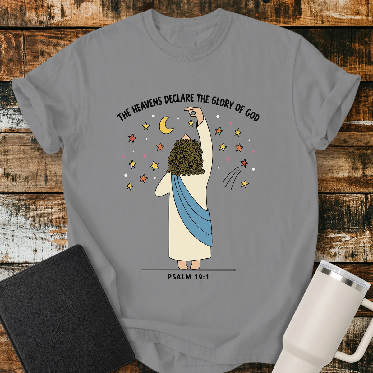 grey Christian t-shirt featuring a whimsical illustration of God placing stars in the heavens, with the text “The Heavens Declare the Glory of God” and Psalm 19:1.