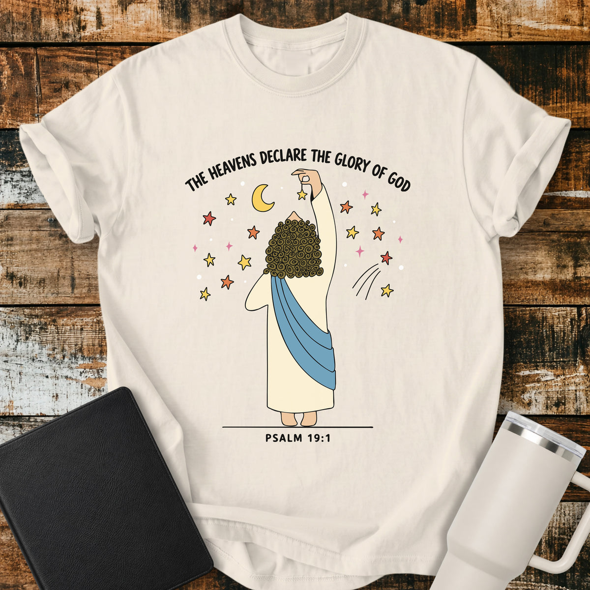ivory Christian t-shirt featuring a whimsical illustration of God placing stars in the heavens, with the text “The Heavens Declare the Glory of God” and Psalm 19:1.