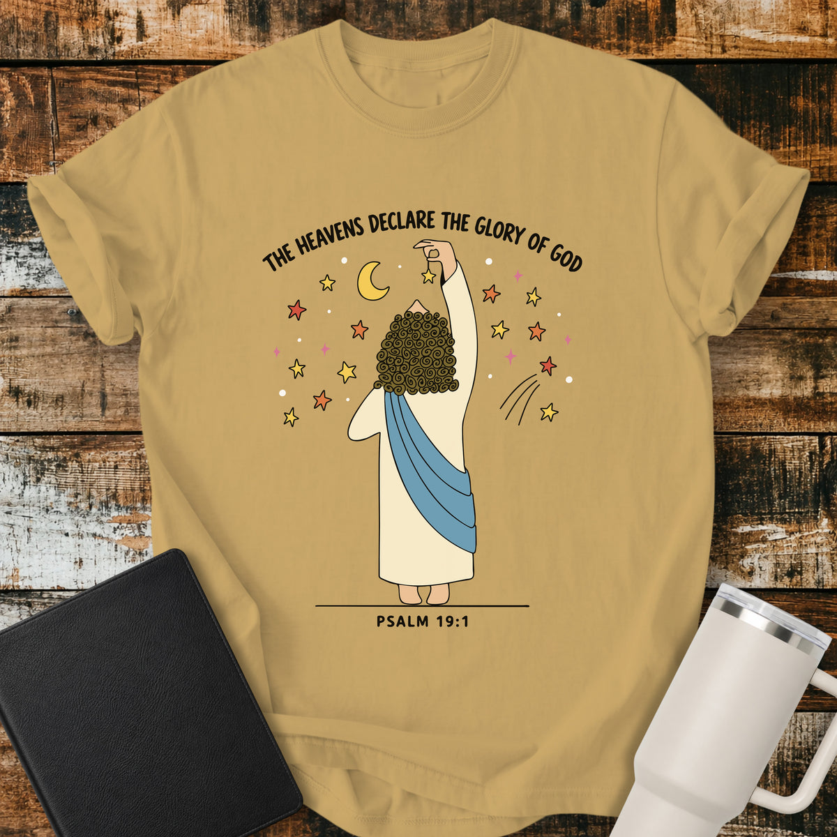 Mustard yellow Christian t-shirt featuring a whimsical illustration of God placing stars in the heavens, with the text “The Heavens Declare the Glory of God” and Psalm 19:1.