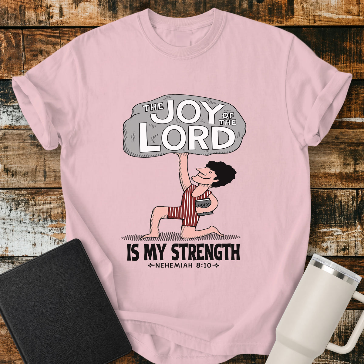 blossom pink Christian t-shirt featuring a vintage-style illustration of a man lifting a large stone reading “The Joy of the Lord,” with the words “Is My Strength” and Nehemiah 8:10 below.