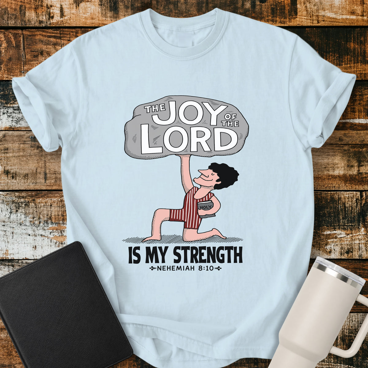 chambray blue Christian t-shirt featuring a vintage-style illustration of a man lifting a large stone reading “The Joy of the Lord,” with the words “Is My Strength” and Nehemiah 8:10 below.