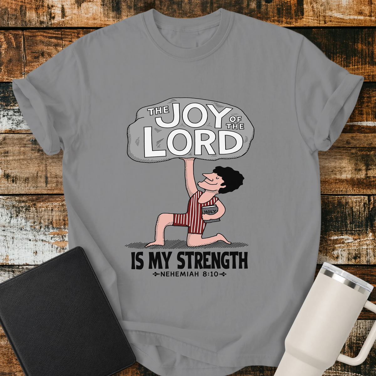 grey Christian t-shirt featuring a vintage-style illustration of a man lifting a large stone reading “The Joy of the Lord,” with the words “Is My Strength” and Nehemiah 8:10 below.