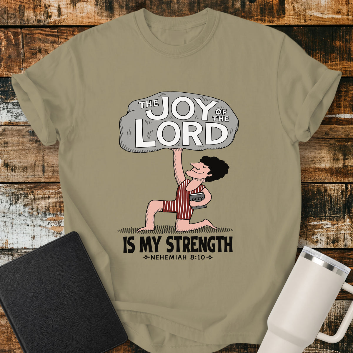 khaki Christian t-shirt featuring a vintage-style illustration of a man lifting a large stone reading “The Joy of the Lord,” with the words “Is My Strength” and Nehemiah 8:10 below.
