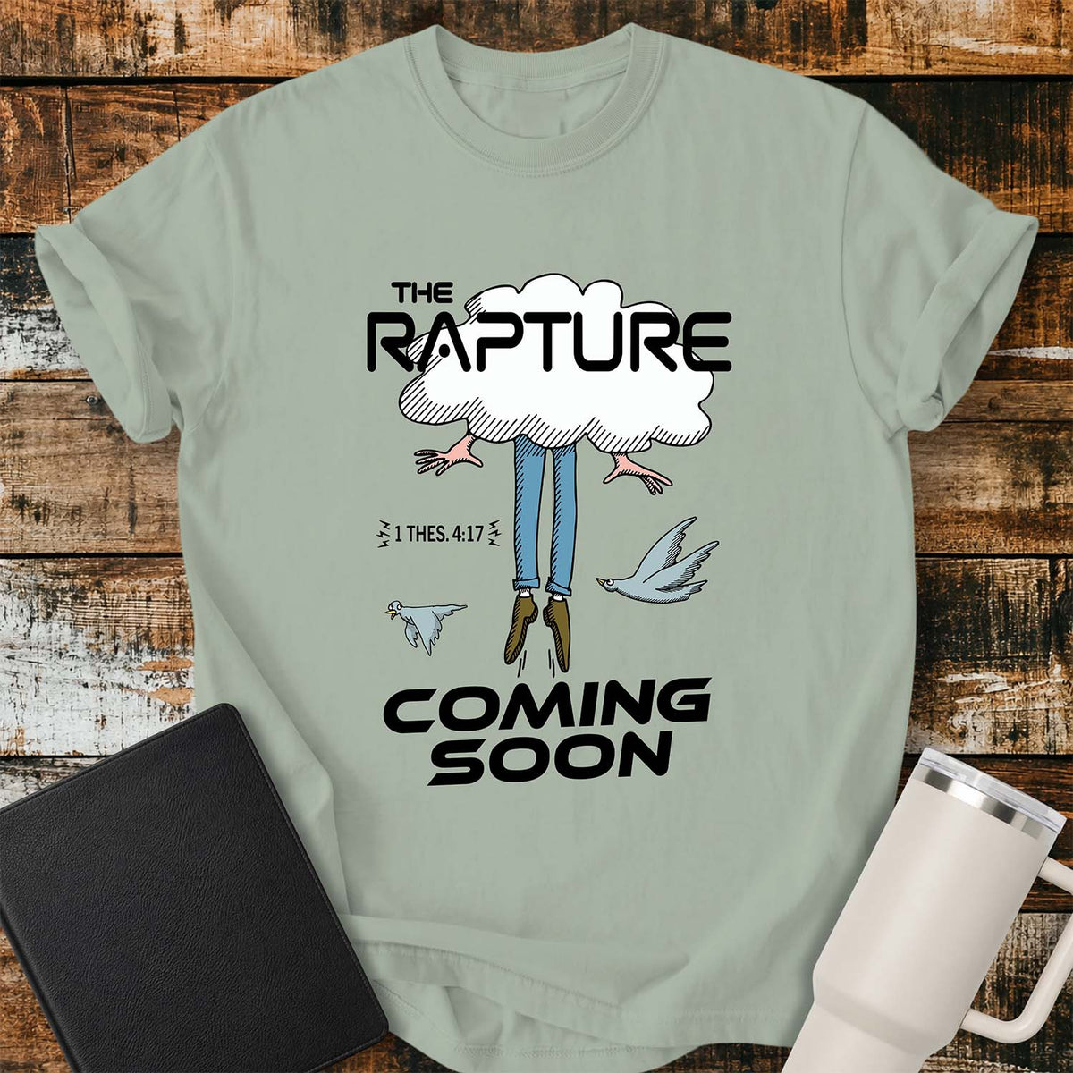 bay green flat-lay t-shirt mockup featuring a humorous illustration of a person being caught up into a cloud, with only their legs and feet visible below. Two birds fly alongside the figure, while bold black lettering reads, "THE RAPTURE" above and "COMING SOON" below. The design includes the Bible reference "1 THES. 4:17." The shirt is displayed on a rustic wood plank background with a black leather Bible and a white insulated travel mug.