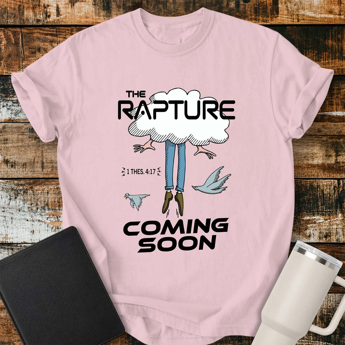 blossom pink flat-lay t-shirt mockup featuring a humorous illustration of a person being caught up into a cloud, with only their legs and feet visible below. Two birds fly alongside the figure, while bold black lettering reads, "THE RAPTURE" above and "COMING SOON" below. The design includes the Bible reference "1 THES. 4:17." The shirt is displayed on a rustic wood plank background with a black leather Bible and a white insulated travel mug.