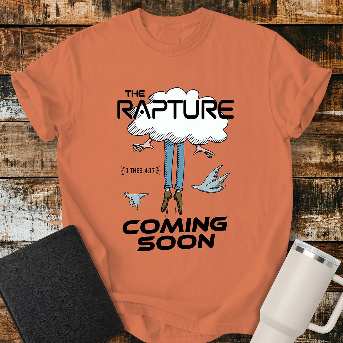 burnt orange flat-lay t-shirt mockup featuring a humorous illustration of a person being caught up into a cloud, with only their legs and feet visible below. Two birds fly alongside the figure, while bold black lettering reads, "THE RAPTURE" above and "COMING SOON" below. The design includes the Bible reference "1 THES. 4:17." The shirt is displayed on a rustic wood plank background with a black leather Bible and a white insulated travel mug.