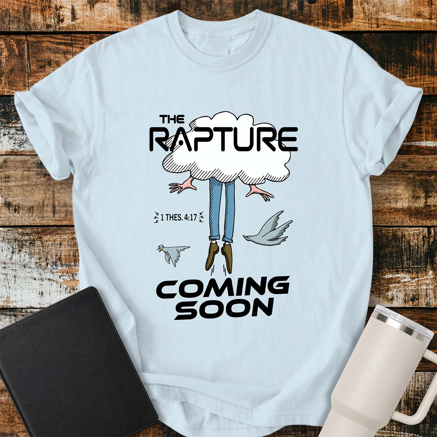 chambray blue flat-lay t-shirt mockup featuring a humorous illustration of a person being caught up into a cloud, with only their legs and feet visible below. Two birds fly alongside the figure, while bold black lettering reads, "THE RAPTURE" above and "COMING SOON" below. The design includes the Bible reference "1 THES. 4:17." The shirt is displayed on a rustic wood plank background with a black leather Bible and a white insulated travel mug.