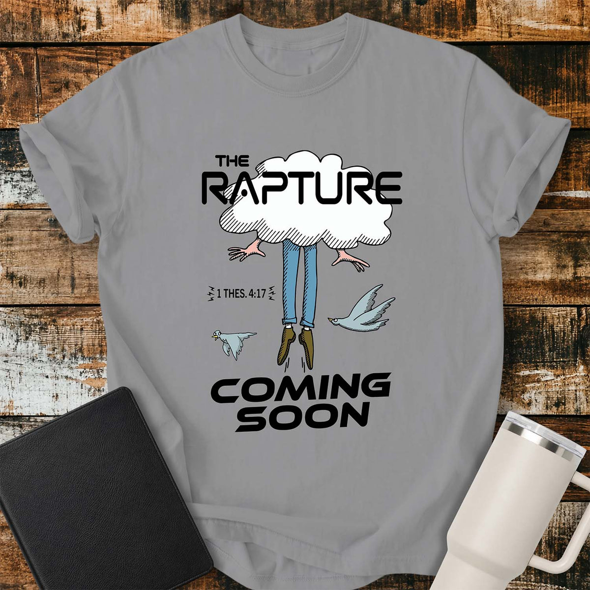 grey flat-lay t-shirt mockup featuring a humorous illustration of a person being caught up into a cloud, with only their legs and feet visible below. Two birds fly alongside the figure, while bold black lettering reads, "THE RAPTURE" above and "COMING SOON" below. The design includes the Bible reference "1 THES. 4:17." The shirt is displayed on a rustic wood plank background with a black leather Bible and a white insulated travel mug.