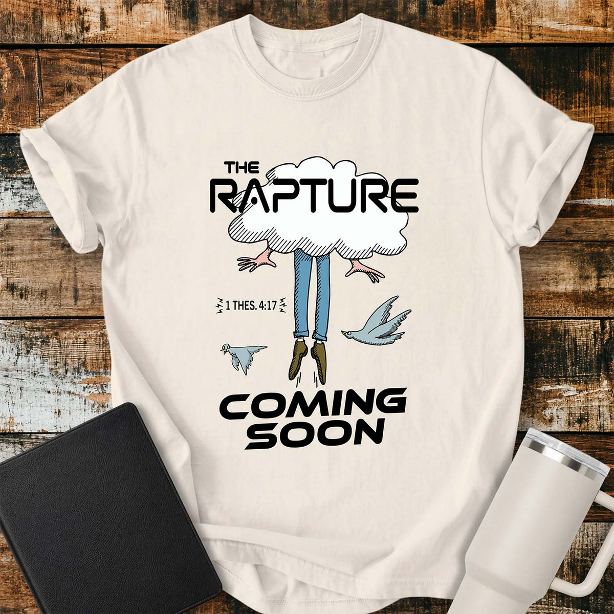 ivory flat-lay t-shirt mockup featuring a humorous illustration of a person being caught up into a cloud, with only their legs and feet visible below. Two birds fly alongside the figure, while bold black lettering reads, "THE RAPTURE" above and "COMING SOON" below. The design includes the Bible reference "1 THES. 4:17." The shirt is displayed on a rustic wood plank background with a black leather Bible and a white insulated travel mug.