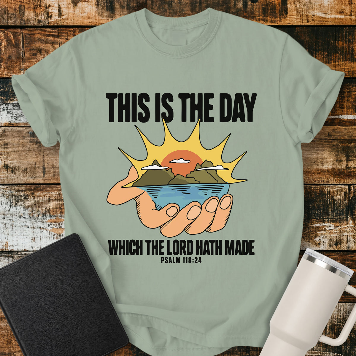 Bay green Christian T-shirt featuring a hand holding a peaceful mountain, water, and sunrise scene with the KJV-inspired message “This Is the Day Which the Lord Hath Made.” Based on Psalm 118:24, this uplifting retro-style design celebrates each day as a gift from God.