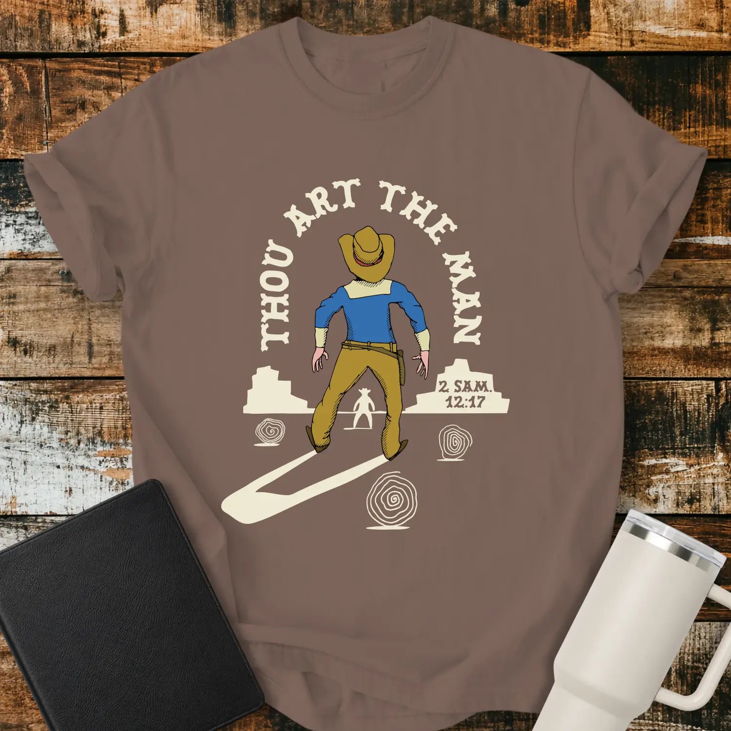 espresso brown graphic T-shirt mockup featuring a western scene with a cowboy facing another figure beneath the phrase “Thou Art the Man” and a 2 Samuel 12:7 reference, styled with a black Bible and white tumbler on a rustic wood background.