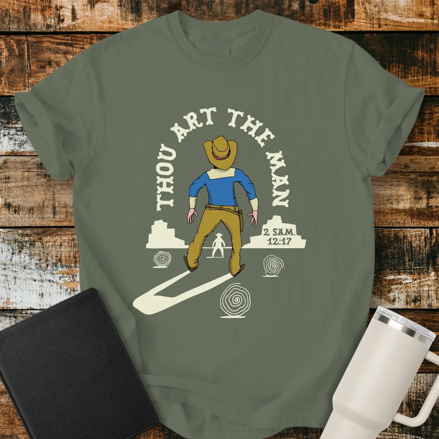 moss green graphic T-shirt mockup featuring a western scene with a cowboy facing another figure beneath the phrase “Thou Art the Man” and a 2 Samuel 12:7 reference, styled with a black Bible and white tumbler on a rustic wood background.