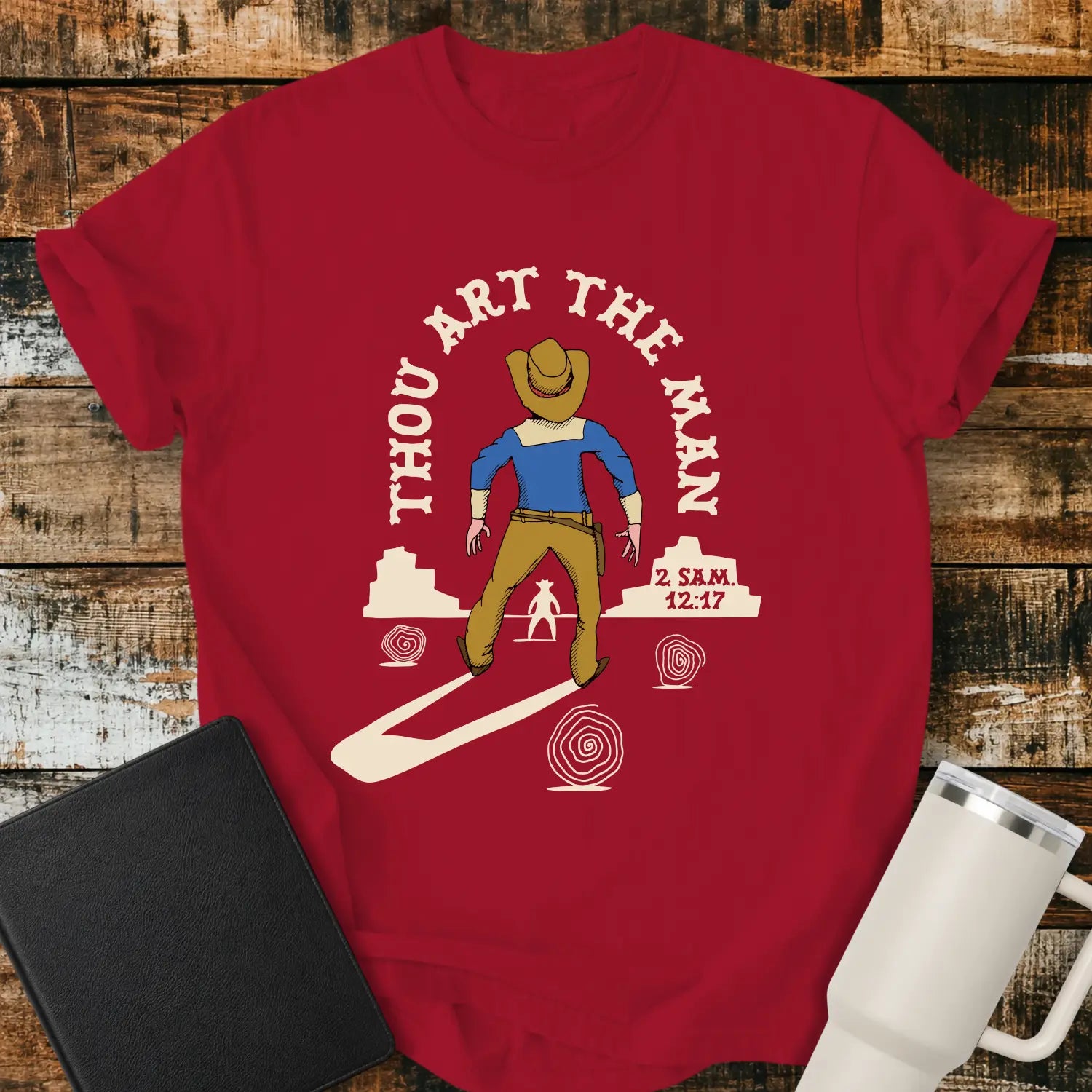 red graphic T-shirt mockup featuring a western scene with a cowboy facing another figure beneath the phrase “Thou Art the Man” and a 2 Samuel 12:7 reference, styled with a black Bible and white tumbler on a rustic wood background.