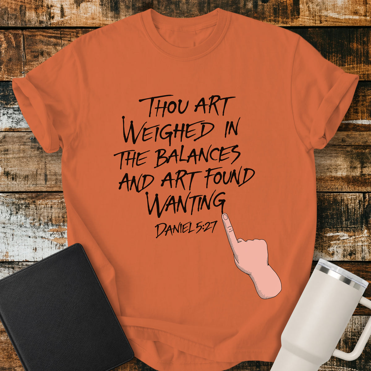 burnt orange Christian t-shirt featuring hand-lettered typography with Daniel 5:27: “Thou art weighed in the balances, and art found wanting,” paired with a pointing-hand illustration.