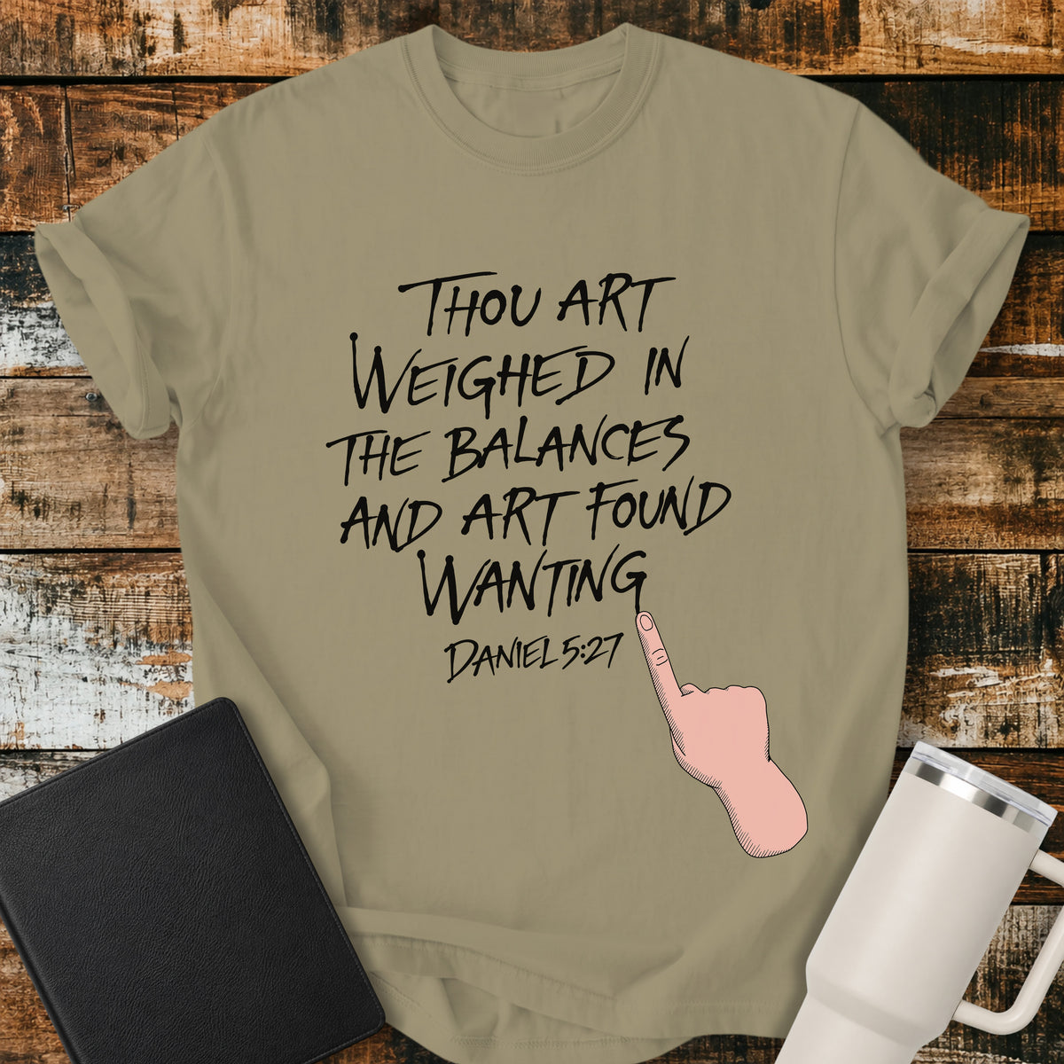 khaki Christian t-shirt featuring hand-lettered typography with Daniel 5:27: “Thou art weighed in the balances, and art found wanting,” paired with a pointing-hand illustration.