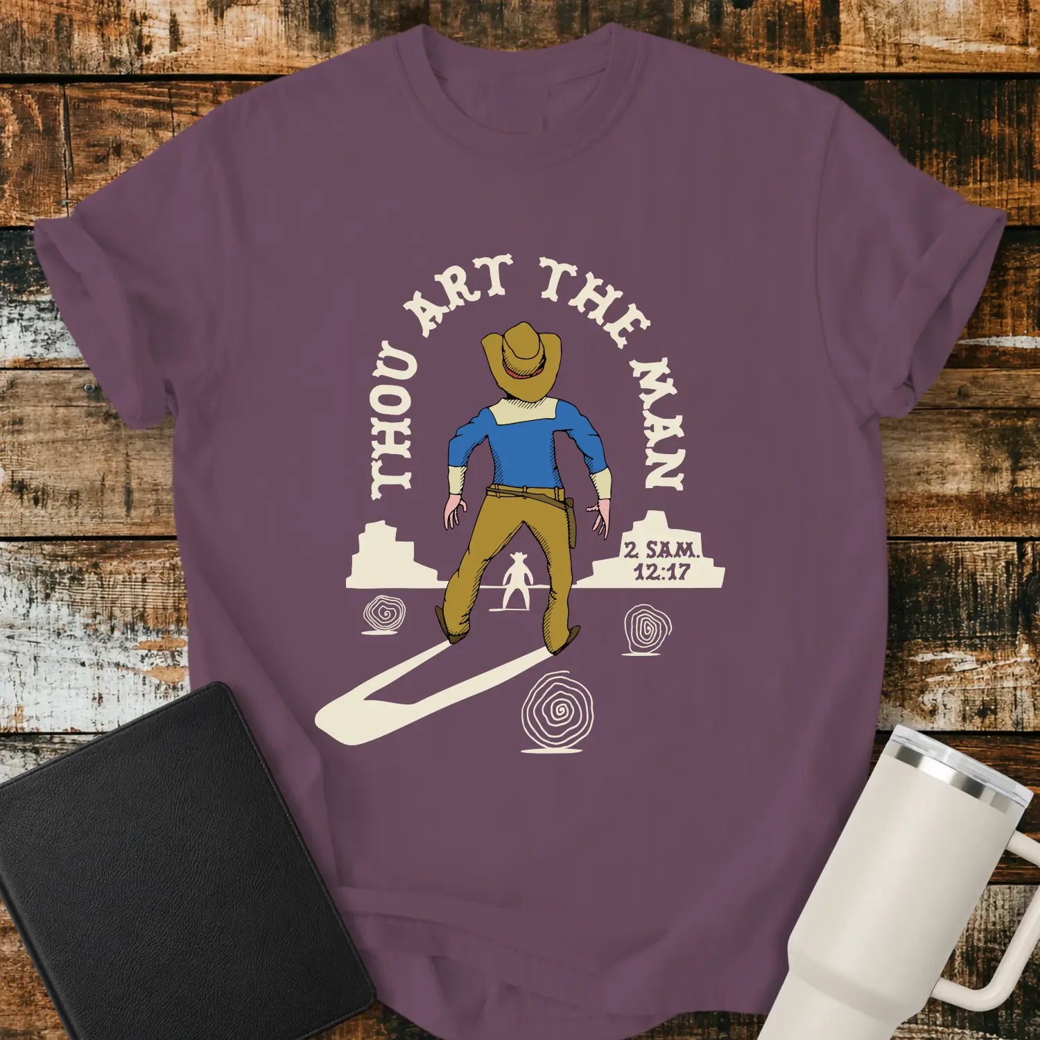 berry graphic T-shirt mockup featuring a western scene with a cowboy facing another figure beneath the phrase “Thou Art the Man” and a 2 Samuel 12:7 reference, styled with a black Bible and white tumbler on a rustic wood background.