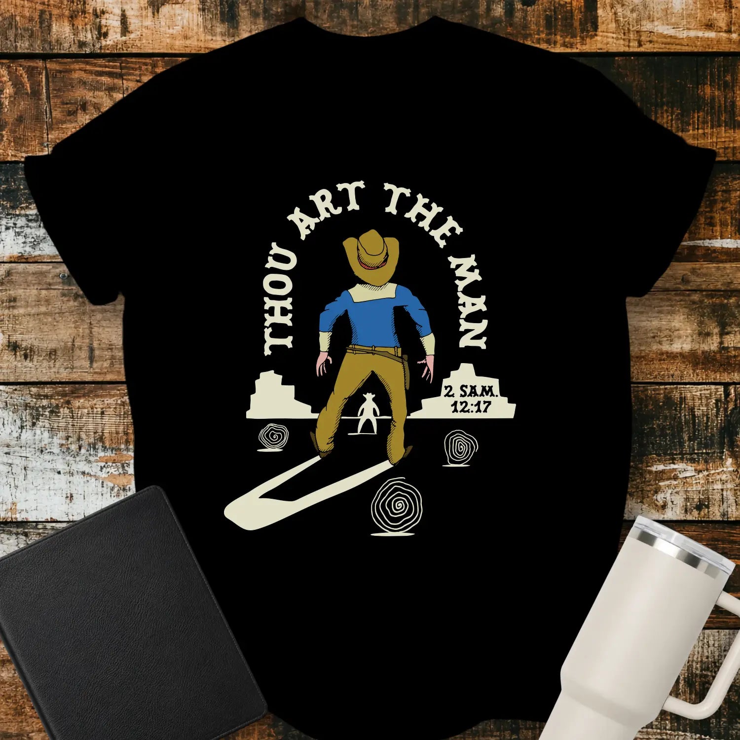 black graphic T-shirt mockup featuring a western scene with a cowboy facing another figure beneath the phrase “Thou Art the Man” and a 2 Samuel 12:7 reference, styled with a black Bible and white tumbler on a rustic wood background.
