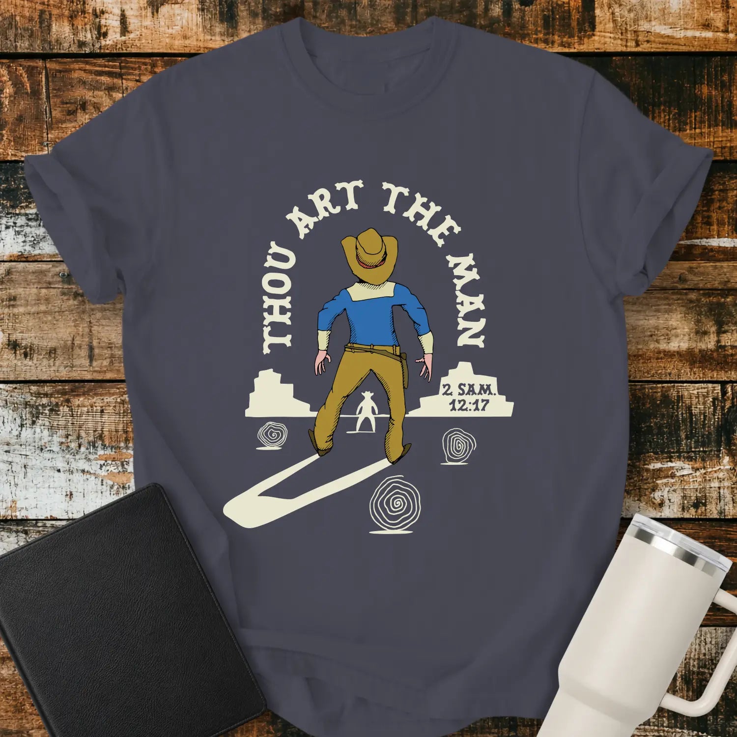 Navy blue graphic T-shirt mockup featuring a western scene with a cowboy facing another figure beneath the phrase “Thou Art the Man” and a 2 Samuel 12:7 reference, styled with a black Bible and white tumbler on a rustic wood background.