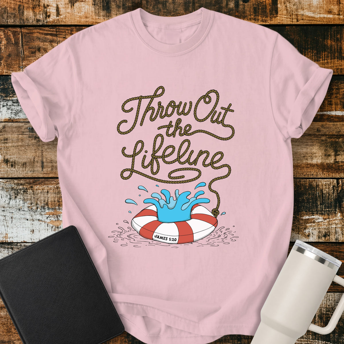blossom pink flat-lay t-shirt mockup featuring rope-style lettering that reads, “THROW OUT THE LIFELINE,” flowing down into a red-and-white life preserver splashing in water. The design includes the Bible reference “James 5:20.” The shirt is displayed on a rustic wood plank background alongside a black leather Bible and a white insulated travel mug.