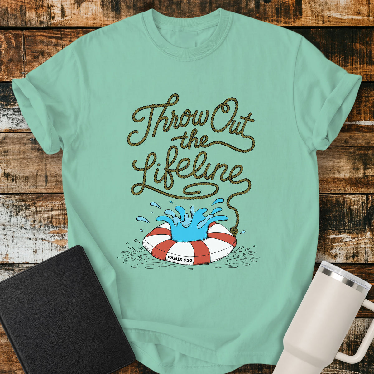 island reef green flat-lay t-shirt mockup featuring rope-style lettering that reads, “THROW OUT THE LIFELINE,” flowing down into a red-and-white life preserver splashing in water. The design includes the Bible reference “James 5:20.” The shirt is displayed on a rustic wood plank background alongside a black leather Bible and a white insulated travel mug.