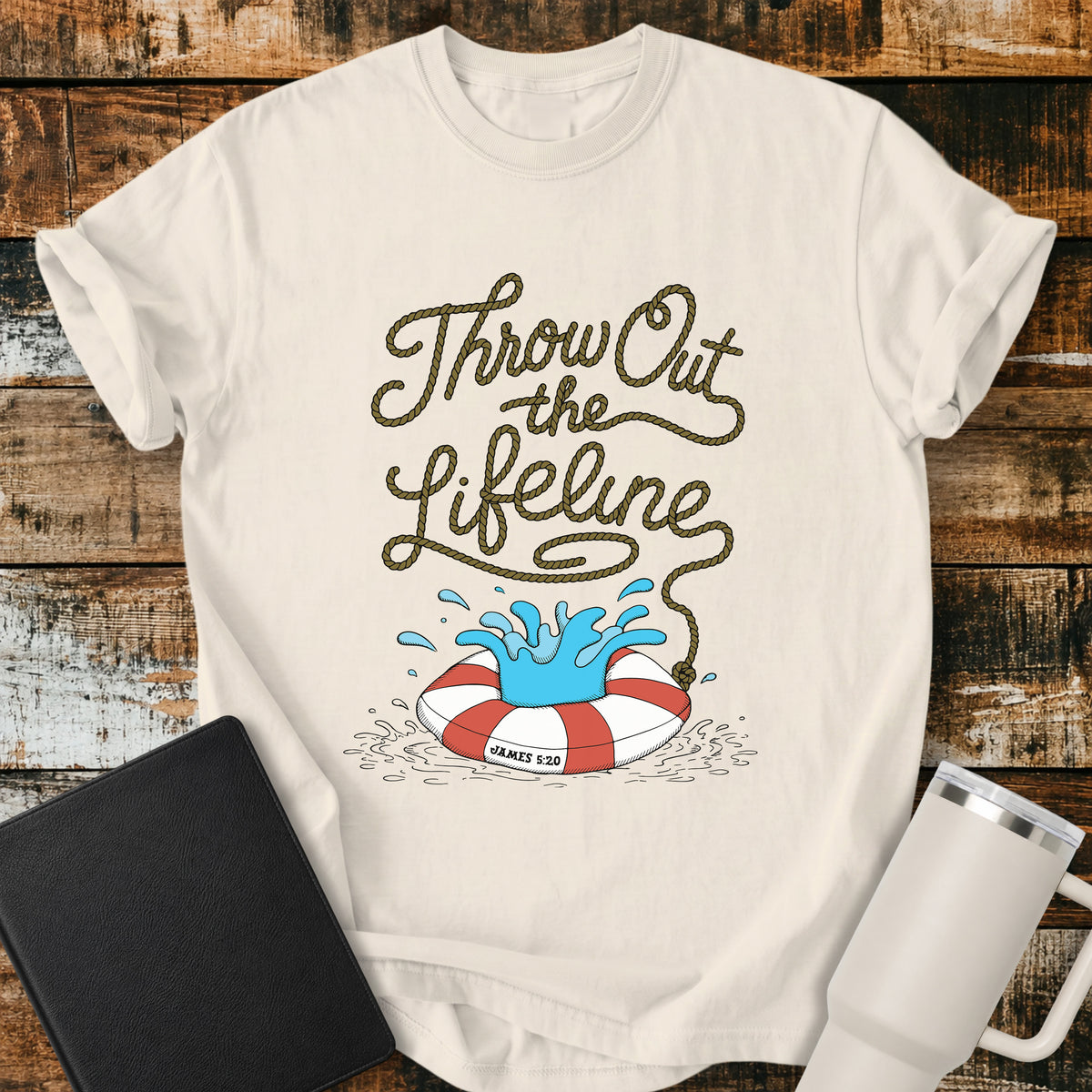 ivory flat-lay t-shirt mockup featuring rope-style lettering that reads, “THROW OUT THE LIFELINE,” flowing down into a red-and-white life preserver splashing in water. The design includes the Bible reference “James 5:20.” The shirt is displayed on a rustic wood plank background alongside a black leather Bible and a white insulated travel mug.