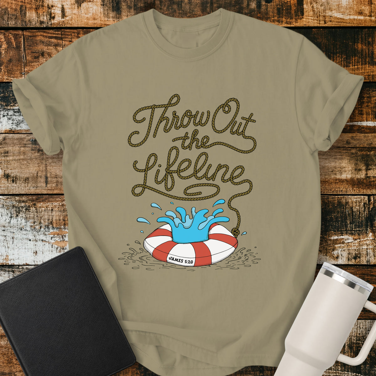 khaki flat-lay t-shirt mockup featuring rope-style lettering that reads, “THROW OUT THE LIFELINE,” flowing down into a red-and-white life preserver splashing in water. The design includes the Bible reference “James 5:20.” The shirt is displayed on a rustic wood plank background alongside a black leather Bible and a white insulated travel mug.