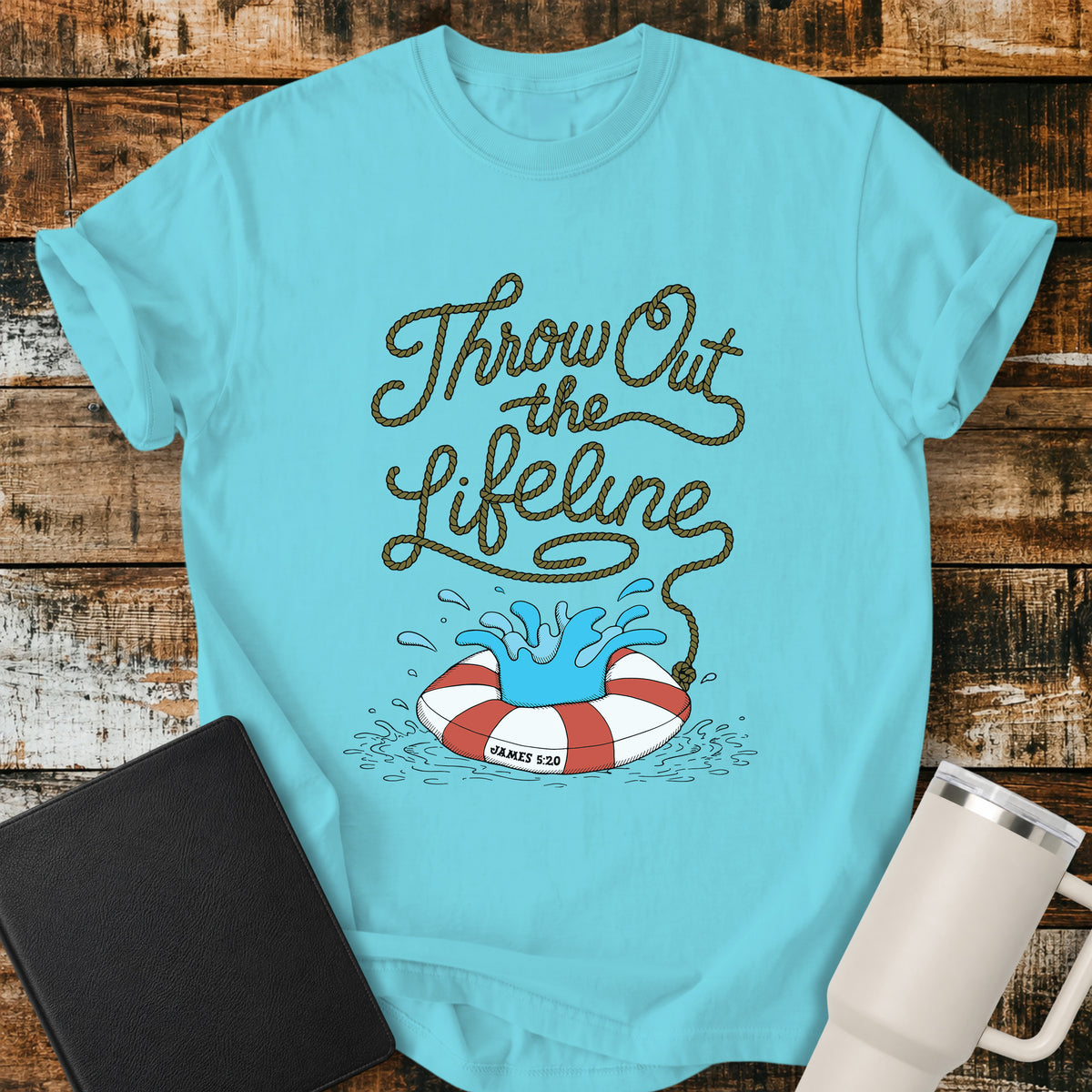 Lagoon blue flat-lay t-shirt mockup featuring rope-style lettering that reads, “THROW OUT THE LIFELINE,” flowing down into a red-and-white life preserver splashing in water. The design includes the Bible reference “James 5:20.” The shirt is displayed on a rustic wood plank background alongside a black leather Bible and a white insulated travel mug.