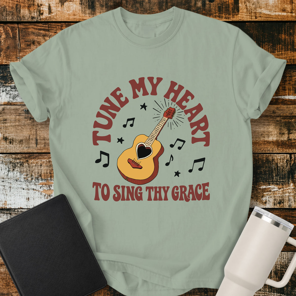 Bay green flat-lay t-shirt mockup featuring a vintage acoustic guitar with a heart-shaped sound hole surrounded by musical notes and stars. Curved retro lettering reads, "TUNE MY HEART" above and "TO SING THY GRACE" below, inspired by the hymn Come Thou Fount of Every Blessing. The shirt is displayed on a rustic wood plank background alongside a black leather Bible and a white insulated travel mug.