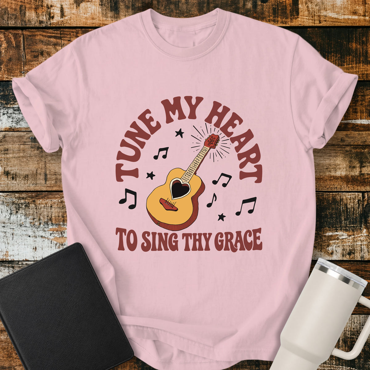 blossom pink flat-lay t-shirt mockup featuring a vintage acoustic guitar with a heart-shaped sound hole surrounded by musical notes and stars. Curved retro lettering reads, "TUNE MY HEART" above and "TO SING THY GRACE" below, inspired by the hymn Come Thou Fount of Every Blessing. The shirt is displayed on a rustic wood plank background alongside a black leather Bible and a white insulated travel mug.