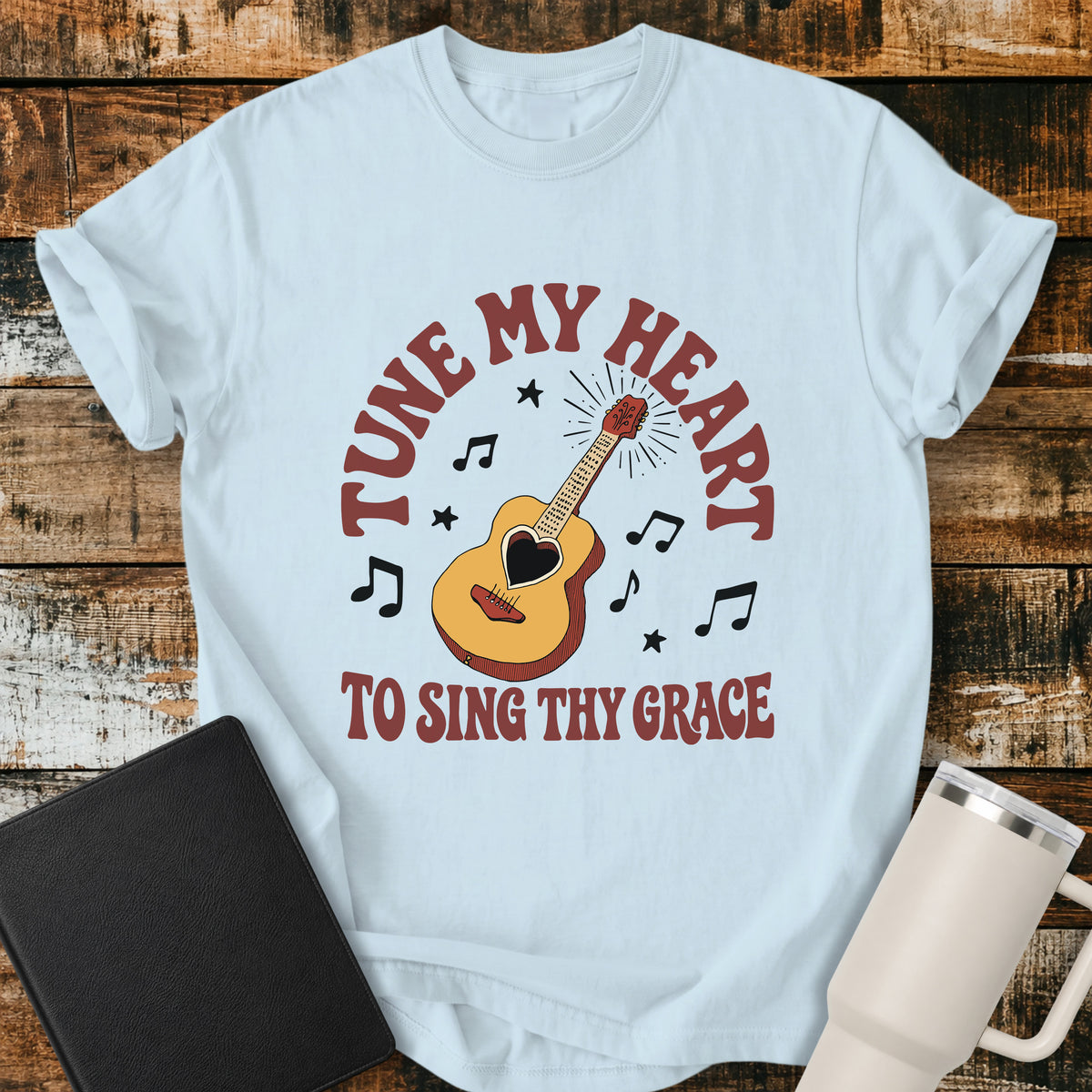 chambray blue flat-lay t-shirt mockup featuring a vintage acoustic guitar with a heart-shaped sound hole surrounded by musical notes and stars. Curved retro lettering reads, "TUNE MY HEART" above and "TO SING THY GRACE" below, inspired by the hymn Come Thou Fount of Every Blessing. The shirt is displayed on a rustic wood plank background alongside a black leather Bible and a white insulated travel mug.