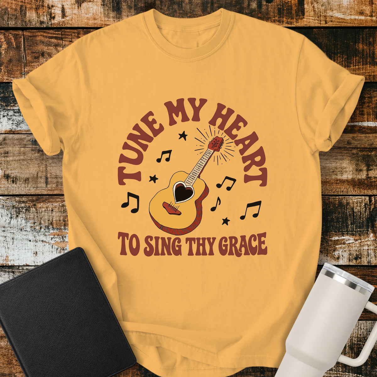 citrus yellow flat-lay t-shirt mockup featuring a vintage acoustic guitar with a heart-shaped sound hole surrounded by musical notes and stars. Curved retro lettering reads, "TUNE MY HEART" above and "TO SING THY GRACE" below, inspired by the hymn Come Thou Fount of Every Blessing. The shirt is displayed on a rustic wood plank background alongside a black leather Bible and a white insulated travel mug.