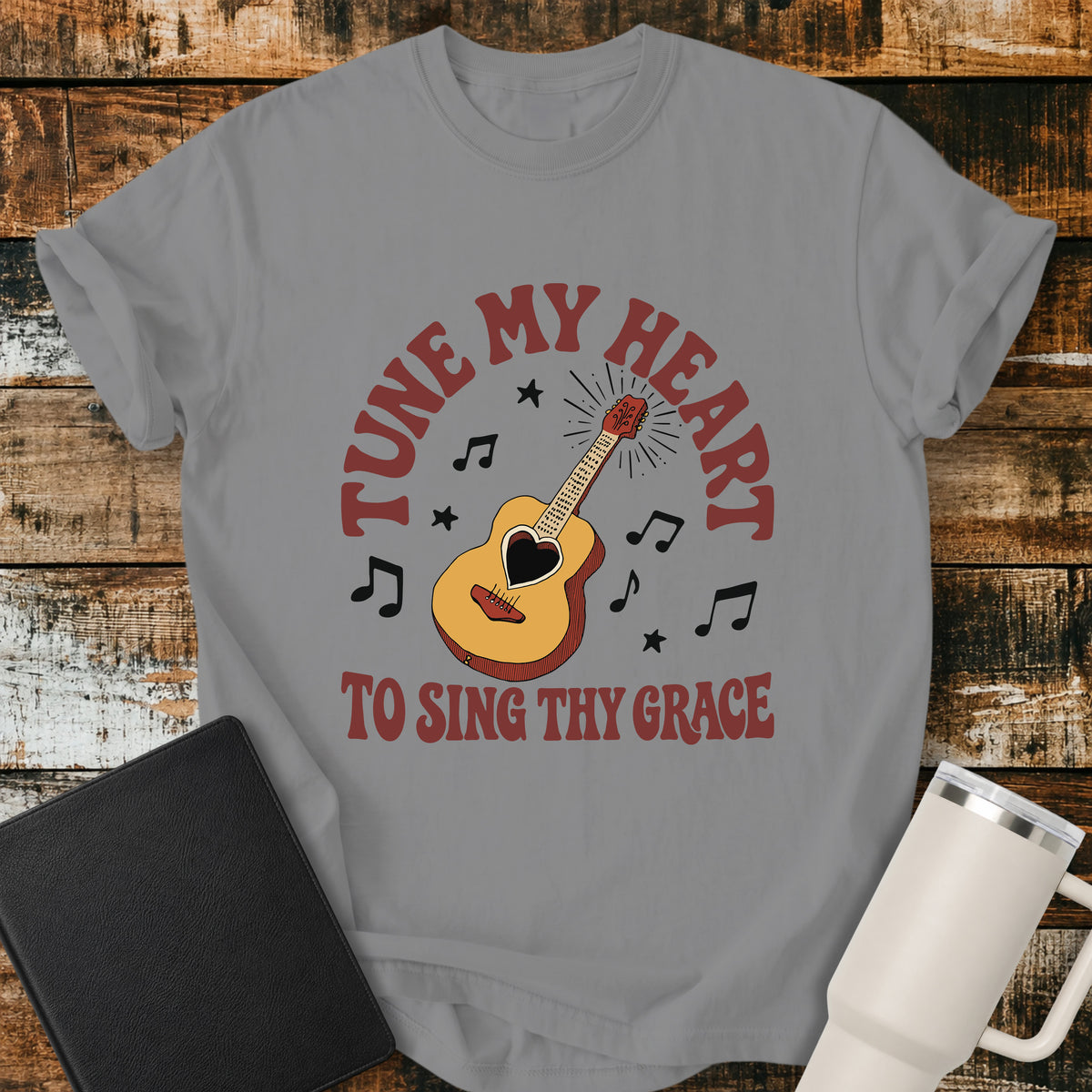 grey flat-lay t-shirt mockup featuring a vintage acoustic guitar with a heart-shaped sound hole surrounded by musical notes and stars. Curved retro lettering reads, "TUNE MY HEART" above and "TO SING THY GRACE" below, inspired by the hymn Come Thou Fount of Every Blessing. The shirt is displayed on a rustic wood plank background alongside a black leather Bible and a white insulated travel mug.