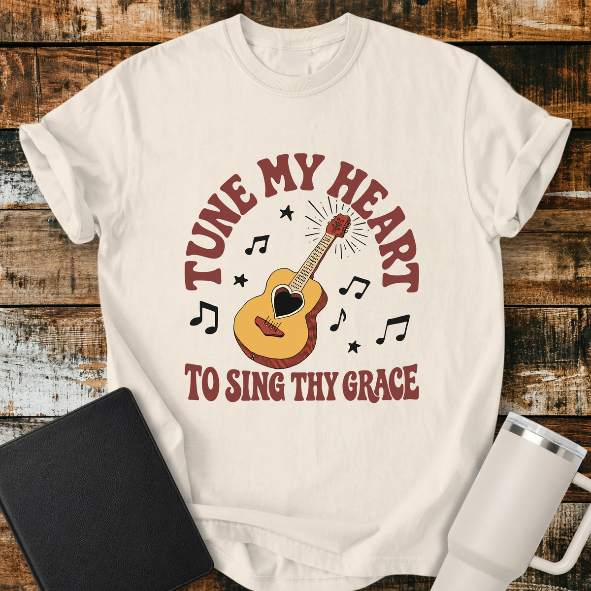 ivory flat-lay t-shirt mockup featuring a vintage acoustic guitar with a heart-shaped sound hole surrounded by musical notes and stars. Curved retro lettering reads, "TUNE MY HEART" above and "TO SING THY GRACE" below, inspired by the hymn Come Thou Fount of Every Blessing. The shirt is displayed on a rustic wood plank background alongside a black leather Bible and a white insulated travel mug.