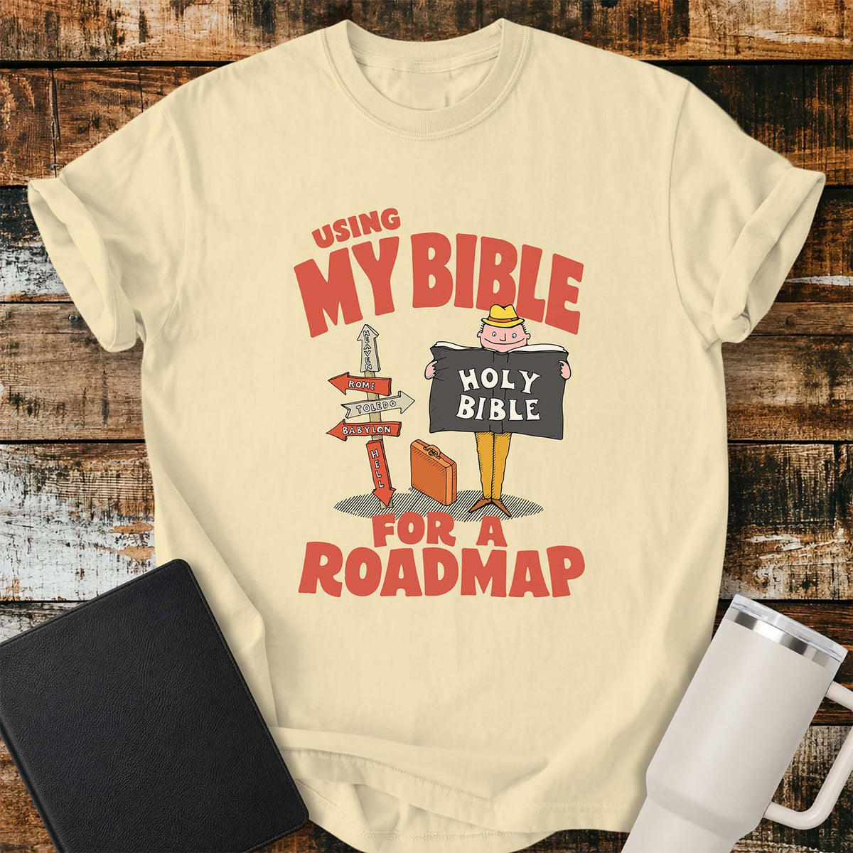 banana yellow T-shirt featuring a retro illustration of a traveler holding an open Holy Bible beside a colorful signpost with directional arrows pointing to places including Heaven, Rome, Toledo, Babylon, and Hell. Large vintage-style coral lettering reads, “USING MY BIBLE FOR A ROADMAP.” The shirt is displayed on a rustic wood plank background with a black Bible on the left and a white insulated tumbler on the right.