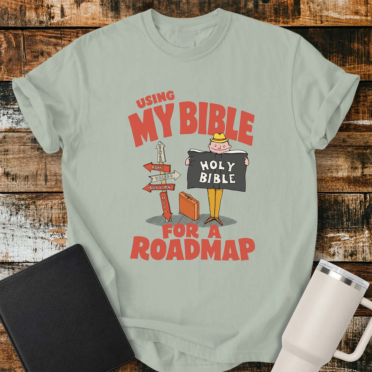bay green T-shirt featuring a retro illustration of a traveler holding an open Holy Bible beside a colorful signpost with directional arrows pointing to places including Heaven, Rome, Toledo, Babylon, and Hell. Large vintage-style coral lettering reads, “USING MY BIBLE FOR A ROADMAP.” The shirt is displayed on a rustic wood plank background with a black Bible on the left and a white insulated tumbler on the right.