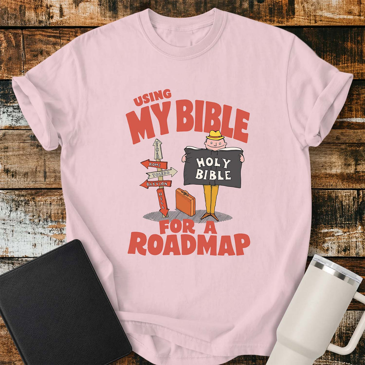 blossom pink T-shirt featuring a retro illustration of a traveler holding an open Holy Bible beside a colorful signpost with directional arrows pointing to places including Heaven, Rome, Toledo, Babylon, and Hell. Large vintage-style coral lettering reads, “USING MY BIBLE FOR A ROADMAP.” The shirt is displayed on a rustic wood plank background with a black Bible on the left and a white insulated tumbler on the right.