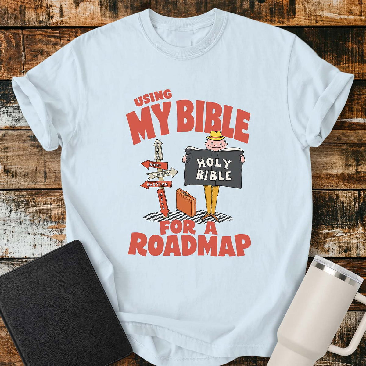 chambray blue T-shirt featuring a retro illustration of a traveler holding an open Holy Bible beside a colorful signpost with directional arrows pointing to places including Heaven, Rome, Toledo, Babylon, and Hell. Large vintage-style coral lettering reads, “USING MY BIBLE FOR A ROADMAP.” The shirt is displayed on a rustic wood plank background with a black Bible on the left and a white insulated tumbler on the right.