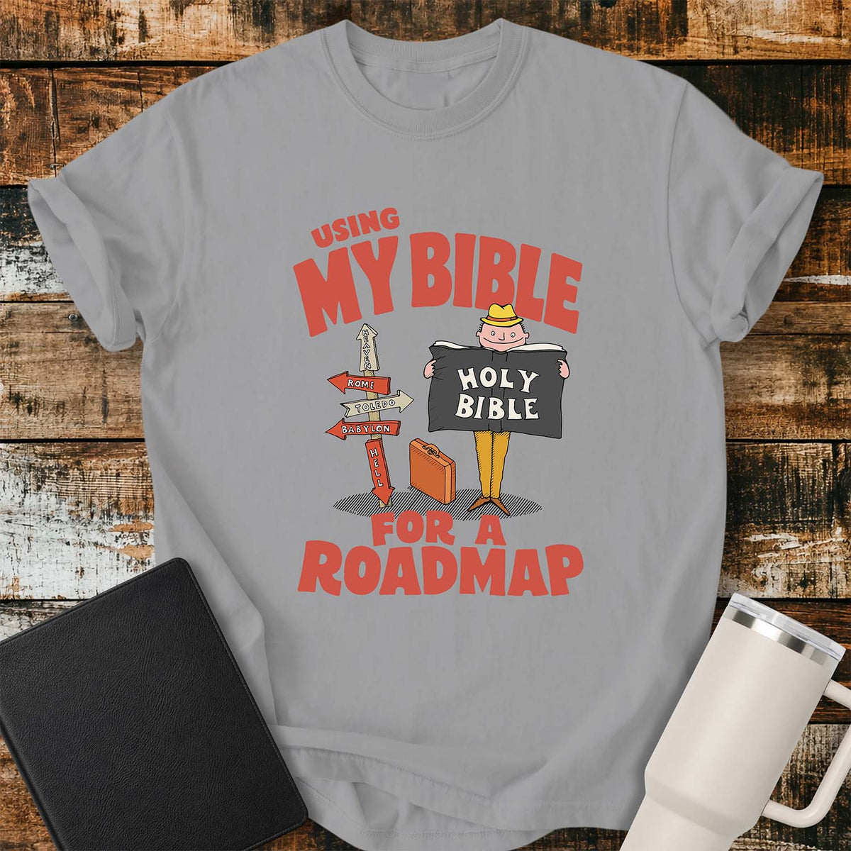 grey T-shirt featuring a retro illustration of a traveler holding an open Holy Bible beside a colorful signpost with directional arrows pointing to places including Heaven, Rome, Toledo, Babylon, and Hell. Large vintage-style coral lettering reads, “USING MY BIBLE FOR A ROADMAP.” The shirt is displayed on a rustic wood plank background with a black Bible on the left and a white insulated tumbler on the right.
