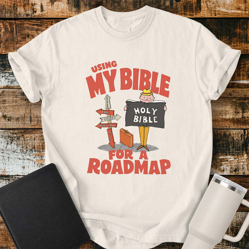 Ivory T-shirt featuring a retro illustration of a traveler holding an open Holy Bible beside a colorful signpost with directional arrows pointing to places including Heaven, Rome, Toledo, Babylon, and Hell. Large vintage-style coral lettering reads, “USING MY BIBLE FOR A ROADMAP.” The shirt is displayed on a rustic wood plank background with a black Bible on the left and a white insulated tumbler on the right.
