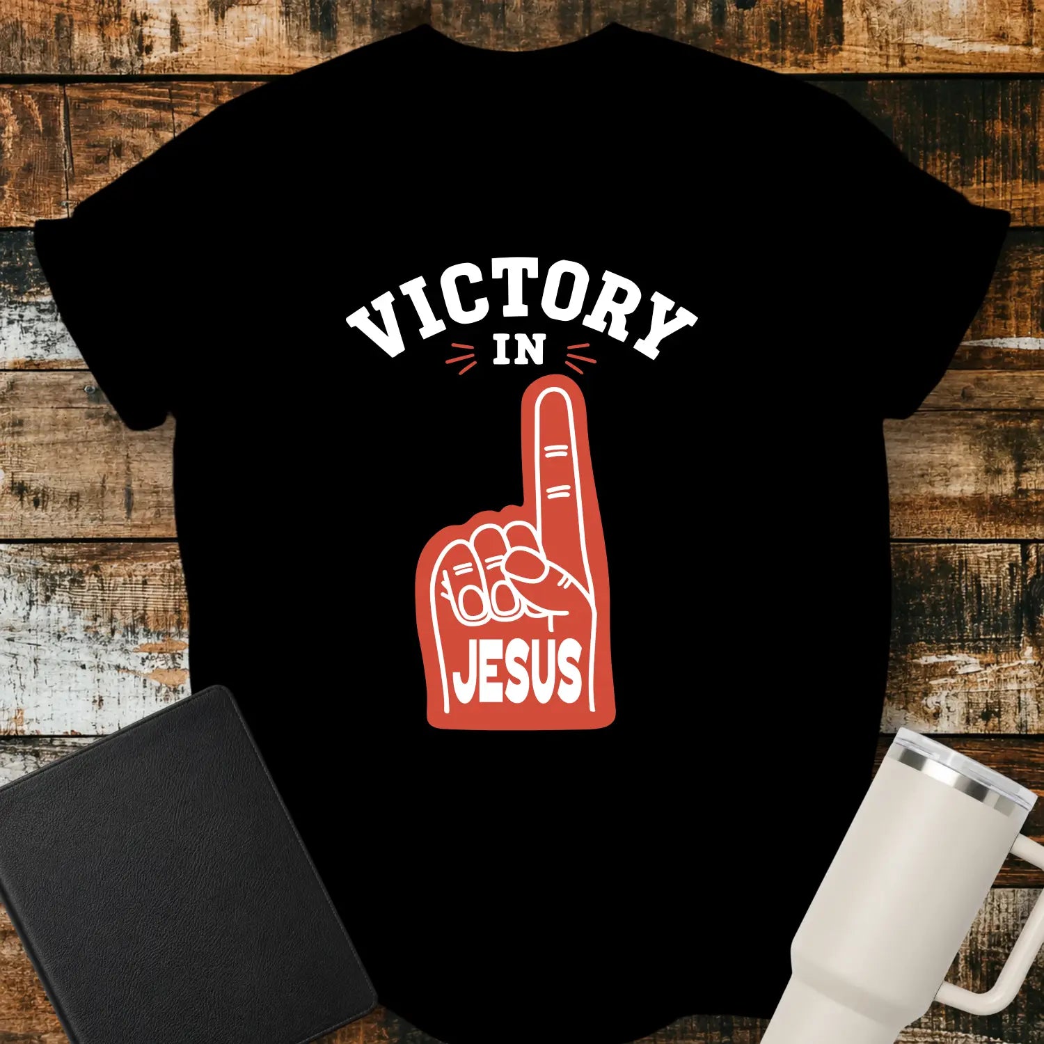black graphic T-shirt mockup featuring a red foam finger with the word “Jesus” and the phrase “Victory in Jesus” in bold white lettering, styled with a black Bible and white tumbler on a rustic wood background.
