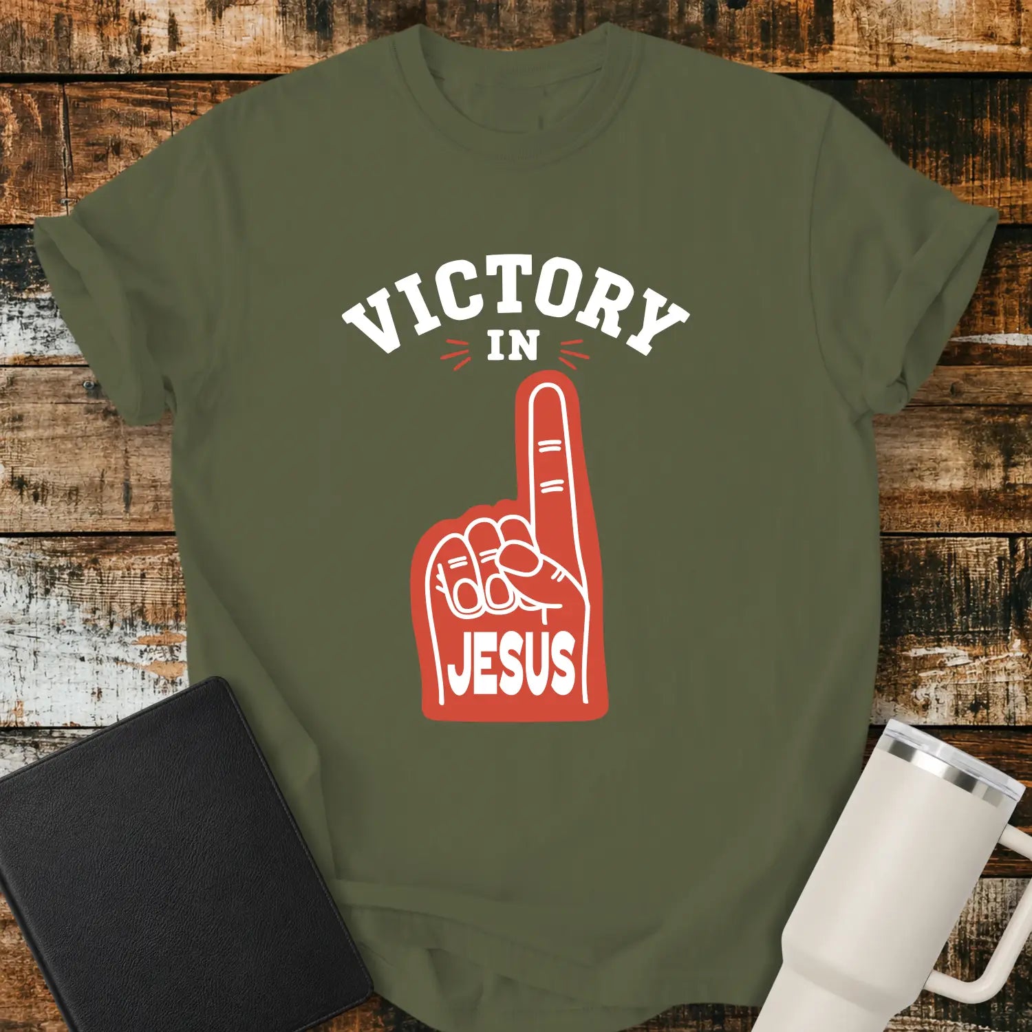 hemp green graphic T-shirt mockup featuring a red foam finger with the word “Jesus” and the phrase “Victory in Jesus” in bold white lettering, styled with a black Bible and white tumbler on a rustic wood background.
