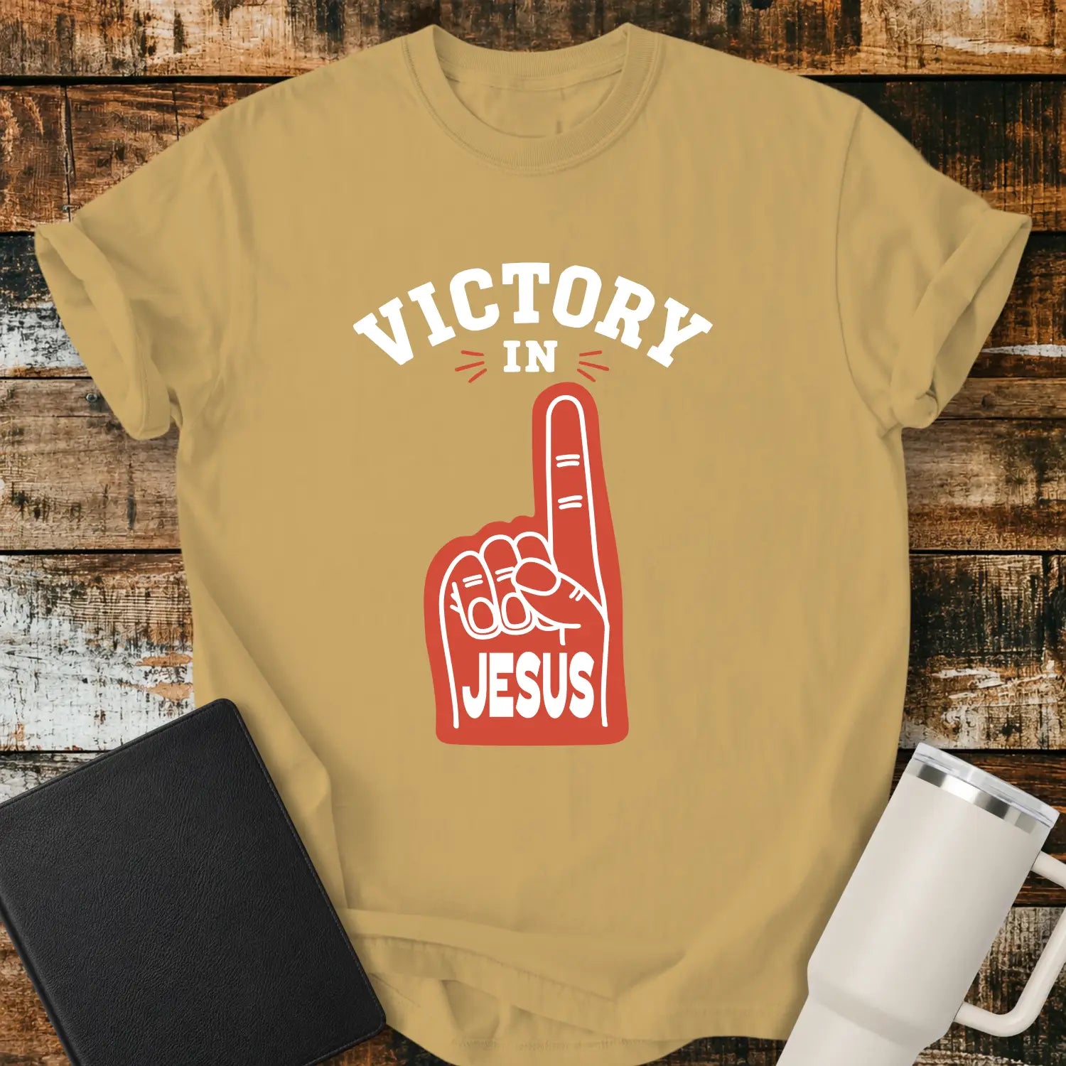 mustard yellow graphic T-shirt mockup featuring a red foam finger with the word “Jesus” and the phrase “Victory in Jesus” in bold white lettering, styled with a black Bible and white tumbler on a rustic wood background.
