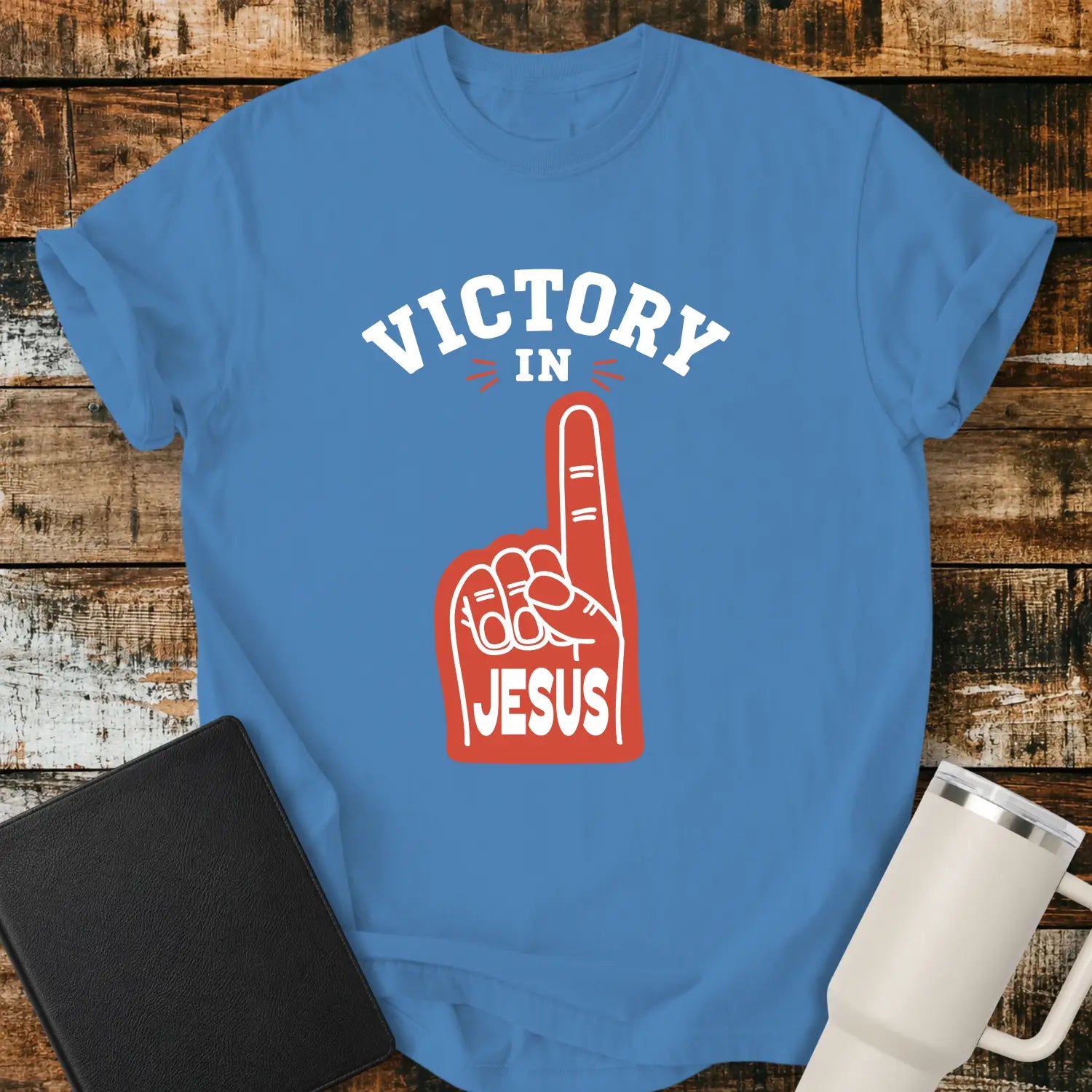 royal caribean blue graphic T-shirt mockup featuring a red foam finger with the word “Jesus” and the phrase “Victory in Jesus” in bold white lettering, styled with a black Bible and white tumbler on a rustic wood background.
