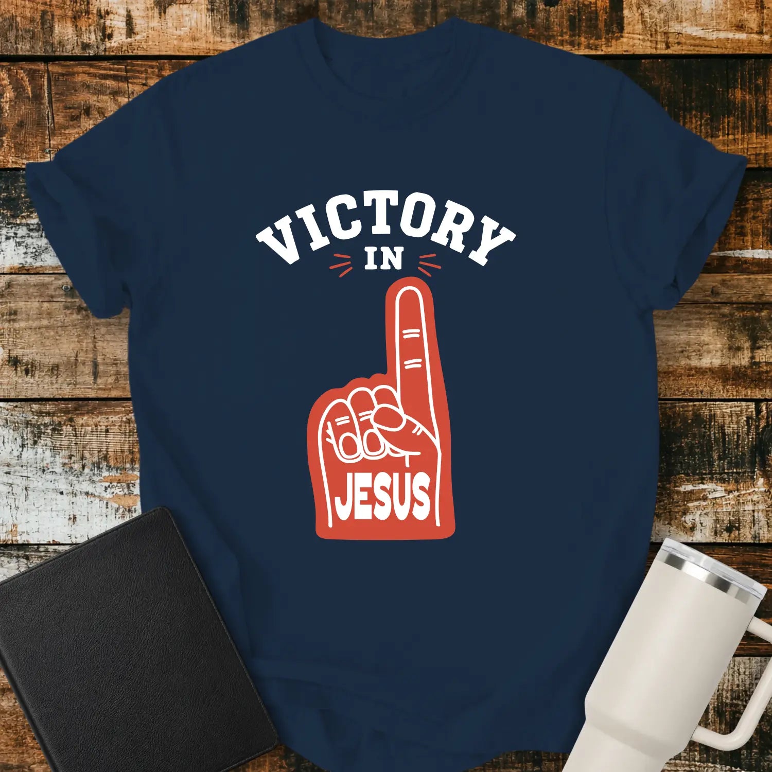 True navy graphic T-shirt mockup featuring a red foam finger with the word “Jesus” and the phrase “Victory in Jesus” in bold white lettering, styled with a black Bible and white tumbler on a rustic wood background.
