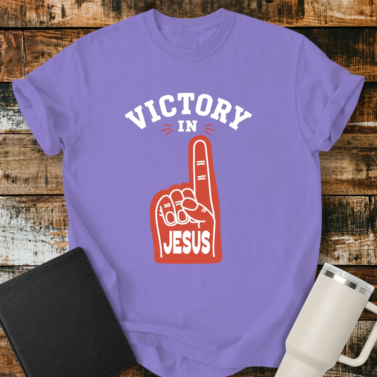 violet graphic T-shirt mockup featuring a red foam finger with the word “Jesus” and the phrase “Victory in Jesus” in bold white lettering, styled with a black Bible and white tumbler on a rustic wood background.
