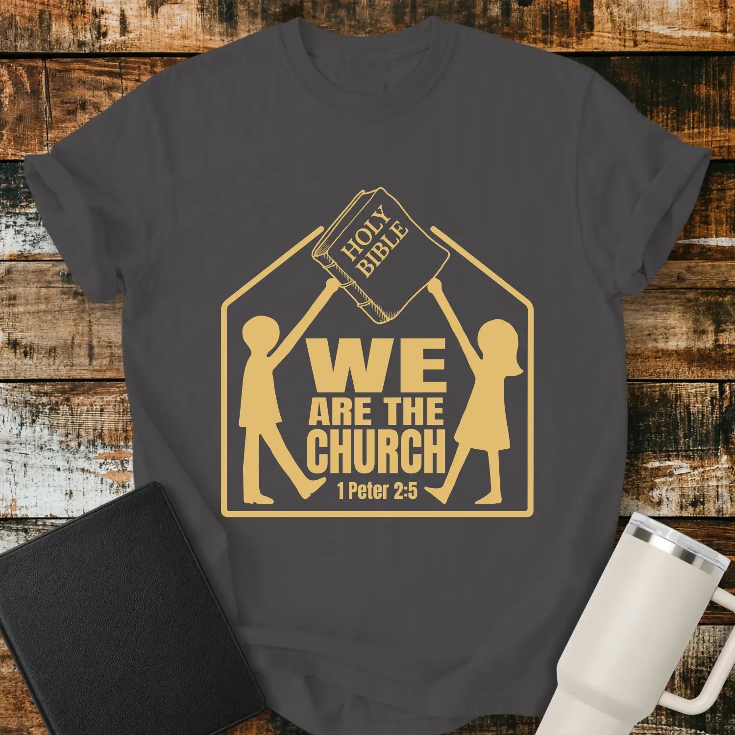 graphite grey graphic T-shirt mockup featuring the phrase “We Are the Church” with a 1 Peter 2:5 reference, showing two figures holding a Holy Bible above a church-shaped outline, styled with a black Bible and white tumbler on a rustic wood background.